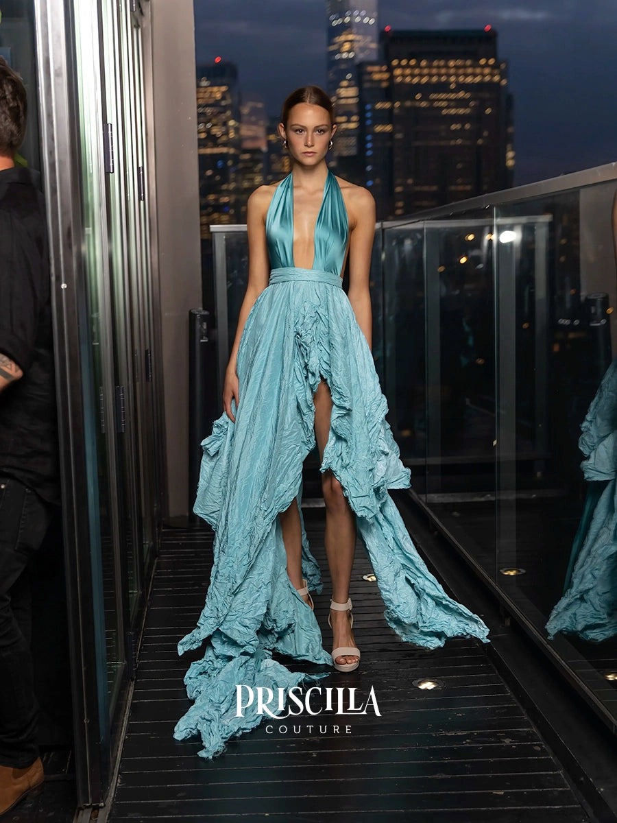 A model walks down a runway wearing a light blue halter gown with a high-low ruffled hem and long train. The background shows a blurred city skyline at dusk, and the brand name 'PRISCILLA COUTURE' is displayed at the bottom.