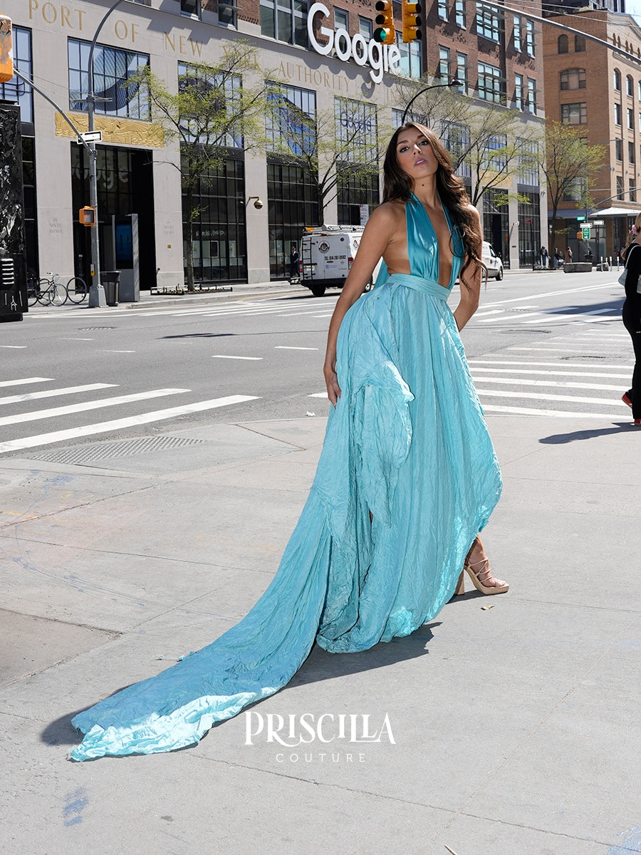 A woman models a light blue halter-neck maxi gown with a dramatic train on a city street. The Priscilla Couture dress features a deep V-neckline, a flowing chiffon-like skirt, and a long train.