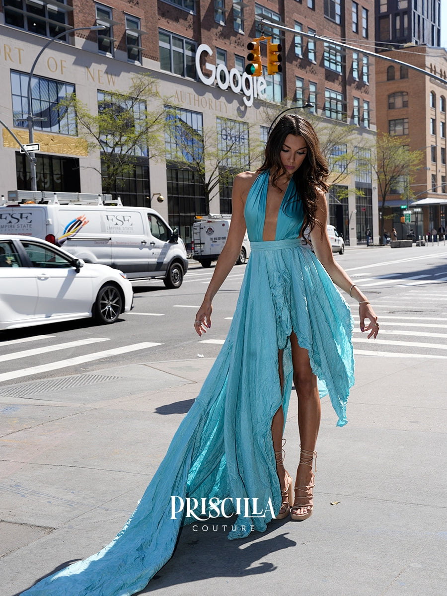 A woman walks on a NYC street wearing a flowing light blue halter gown with a deep V-neckline, high slit, and long train. The Priscilla Couture logo is visible at the bottom of the image.