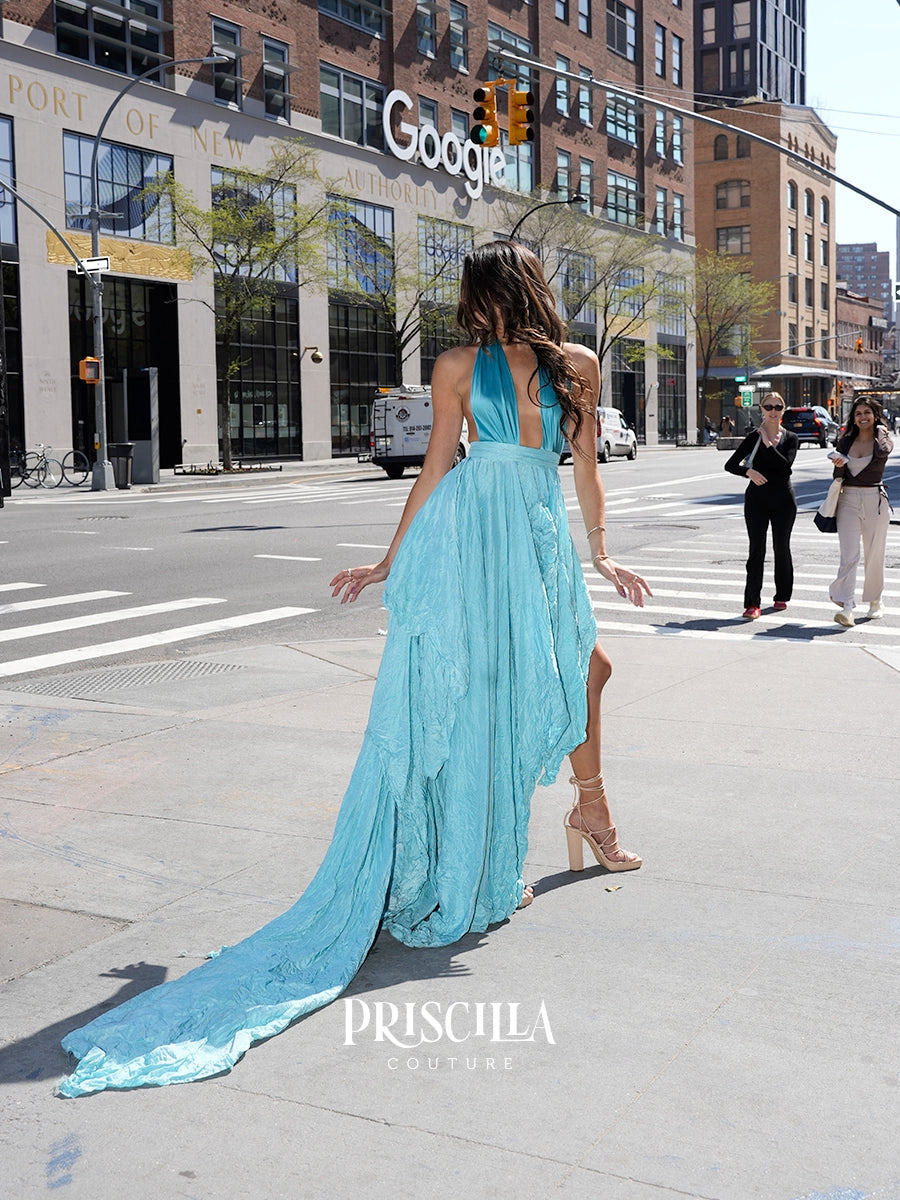 A woman wearing a flowing light blue Priscilla Couture maxi dress with a long train and nude high heels, photographed from behind on a NYC street. The dress has a halter-style bodice and a dramatic cascading skirt.