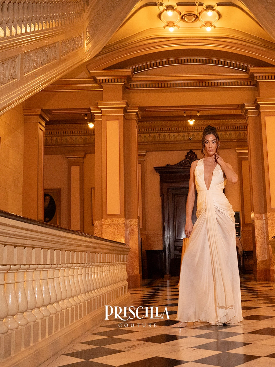A woman models a white Priscilla Couture Hollywood-inspired wedding gown in a grand, warm-toned interior hallway with columns, chandeliers, and a checkered floor.