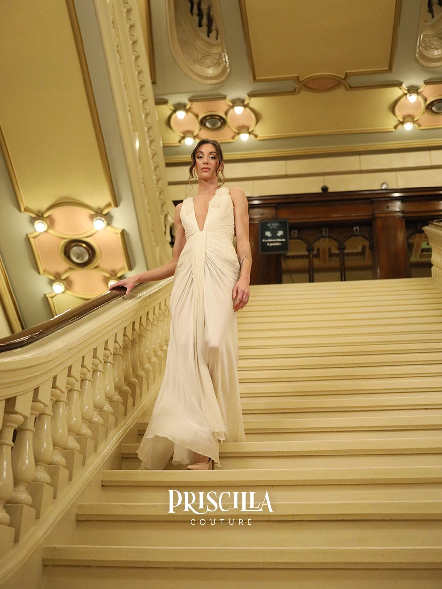 A model wearing a floor-length cream draped Hollywood-inspired wedding gown walks down a grand staircase in a luxurious interior. The brand name 'PRISCILLA COUTURE' is displayed at the bottom of the image.