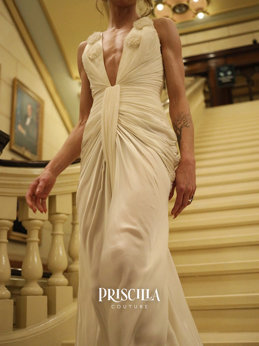 A woman wearing a champagne-colored wedding gown with a deep plunging neckline and gathered draping details, walking down an elegant indoor staircase. The brand name 'PRISCILLA COUTURE' is watermarked at the bottom.