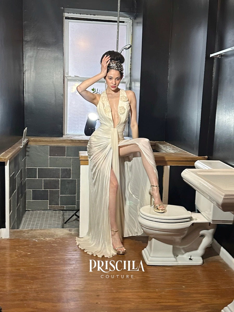 A model wearing a Priscilla Couture champagne-colored Hollywood-inspired silhouette with a high slit and deep plunging neckline, posing in a dark, rustic bathroom setting with a toilet and wooden floor.
