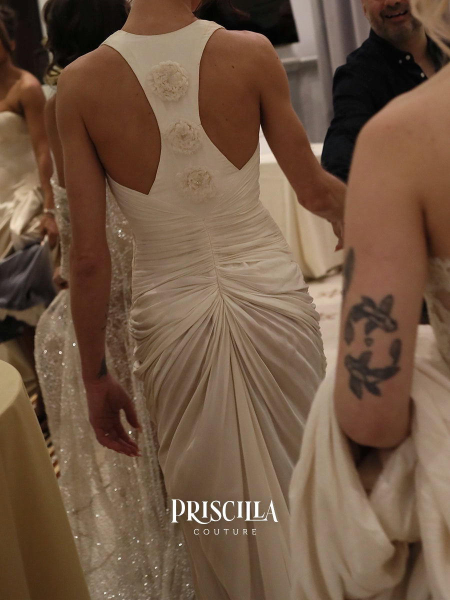 A woman wearing a Priscilla Couture champagne-colored wedding gown, shown from the back in an indoor event setting. The dress features a draped, gathered back with three floral appliques and a deep open back.