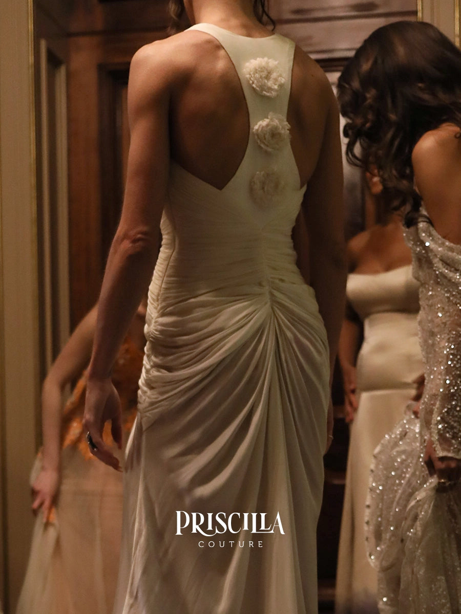 A woman viewed from behind wearing a cream bridal Hollywood-inspired silhouette gown with a draped back, floral applique, and gathered skirt. Another model in a sparkling silver dress stands beside her in a warm indoor setting.