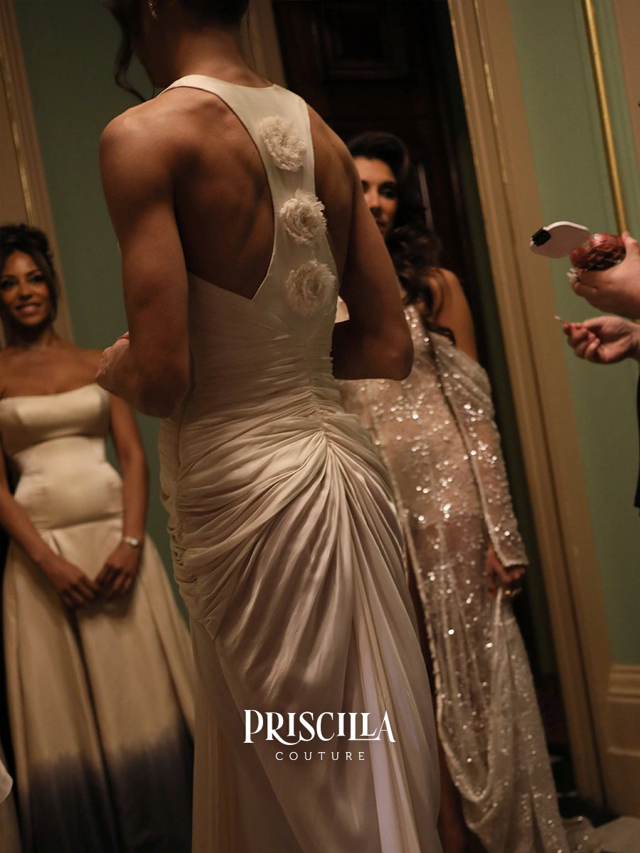 A woman wearing a champagne-colored backless gown with floral appliqués on the back, shown from behind in a mirror reflection inside a dressing room. Other women in formalwear are visible in the background, and a 'PRISCILLA COUTURE' watermark is at the bottom.