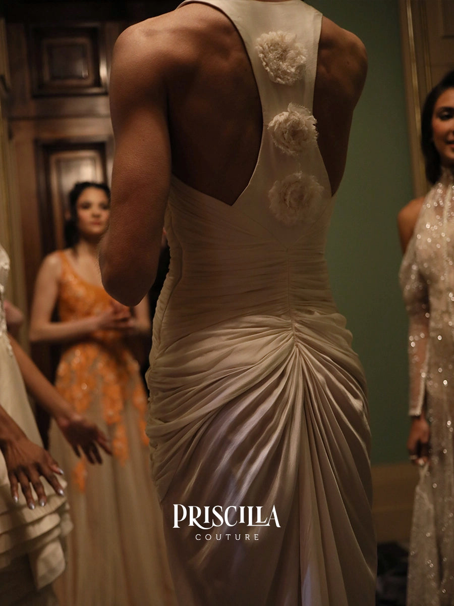 A model from behind wearing a Priscilla Couture champagne-colored Hollywood-inspired silhouette with a draped back and floral embellishments, with other models in formal wear blurred in the background.