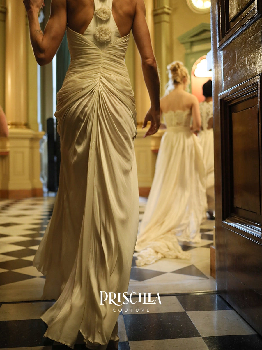 A woman wearing a champagne-colored draped Hollywood-inspired wedding gown walks through an elegant hallway with a black-and-white checkered floor. Another bride in a white gown is visible in the background.