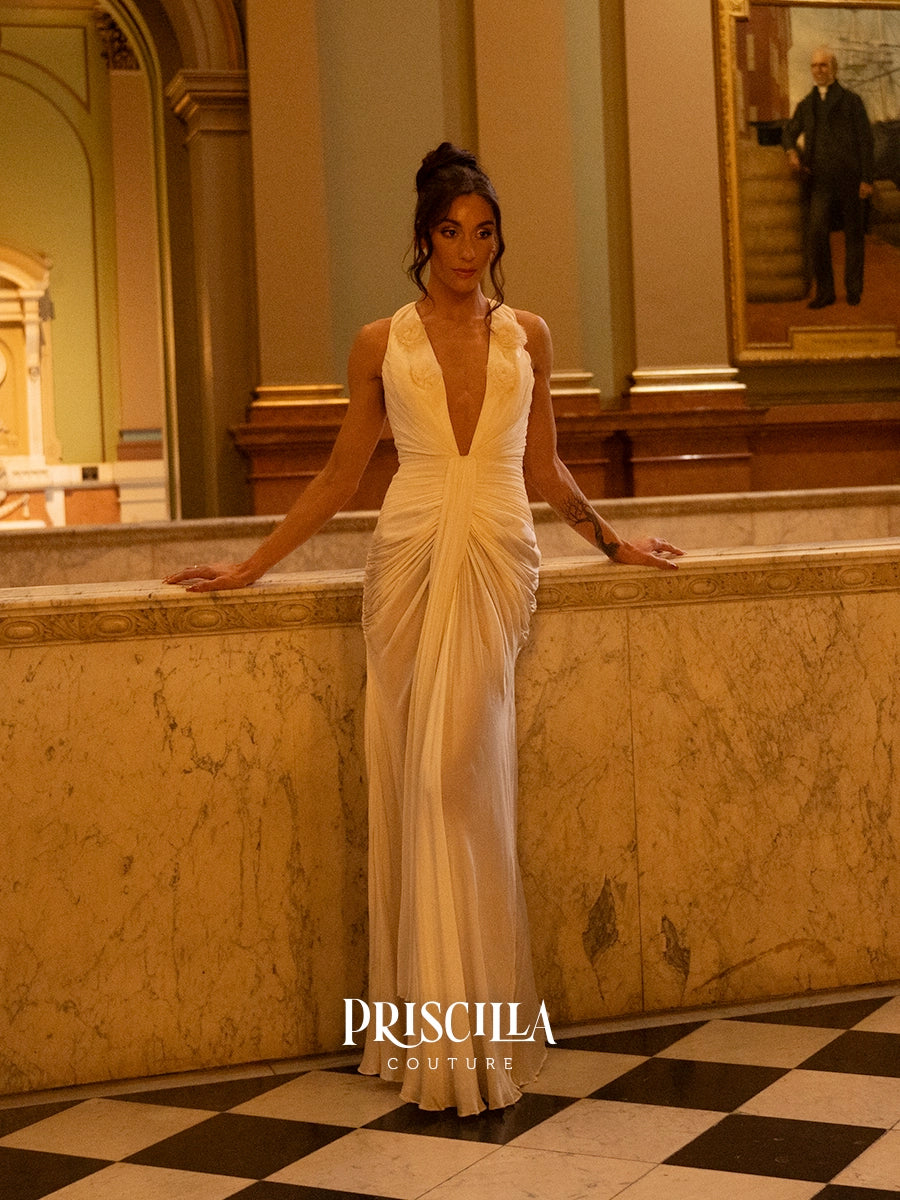 A woman wearing a floor-length Hollywood-inspired white wedding gown with a deep V-neckline and gathered ruching stands indoors against a marble ledge in a grand hall. The PRISCILLA COUTURE logo appears at the bottom.