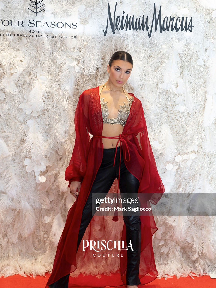 A model stands on a red carpet wearing a sheer red open-front robe over a silver embellished bralette and black pants, posing in front of a white feathered backdrop with event branding.