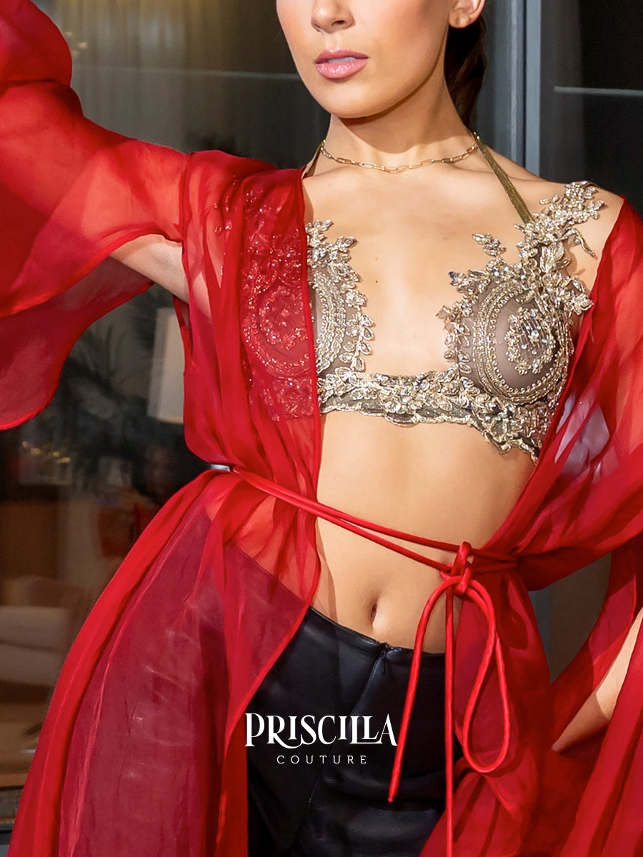 A close-up of a model wearing a red sheer wrap robe tied at the waist over a heavily embellished silver and gold embroidered bralette, with black bottoms. The brand name 'PRISCILLA COUTURE' is watermarked at the bottom.