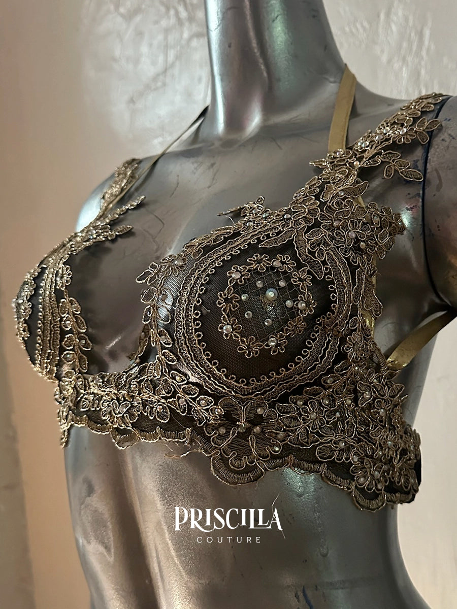 An ornate black Priscilla Couture lace bralette with intricate metallic bronze embroidery and beadwork, displayed on a metallic gray mannequin torso. 