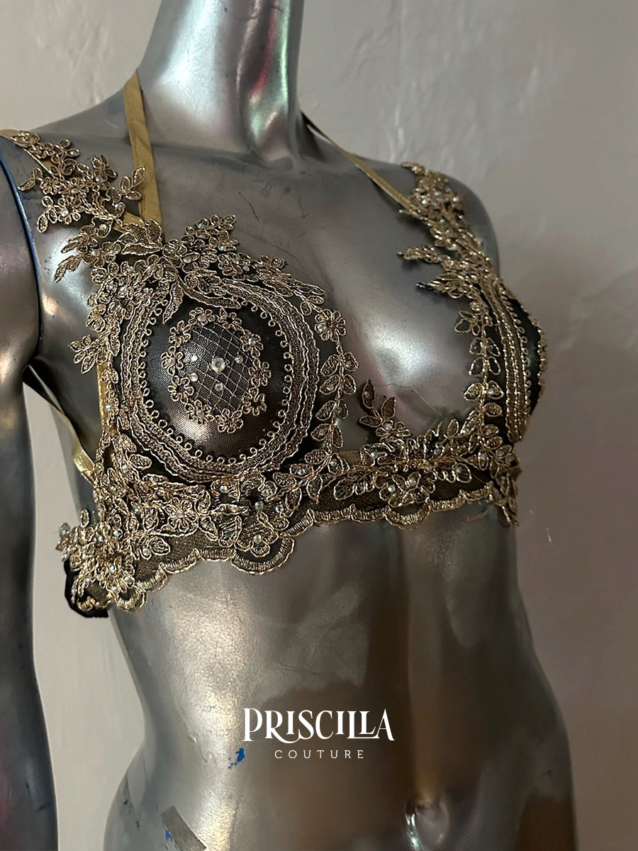 An ornate black and metallic silver Priscilla Couture lace bralette displayed on a metallic mannequin torso. The intricate lace features circular floral patterns and sheer mesh details.
