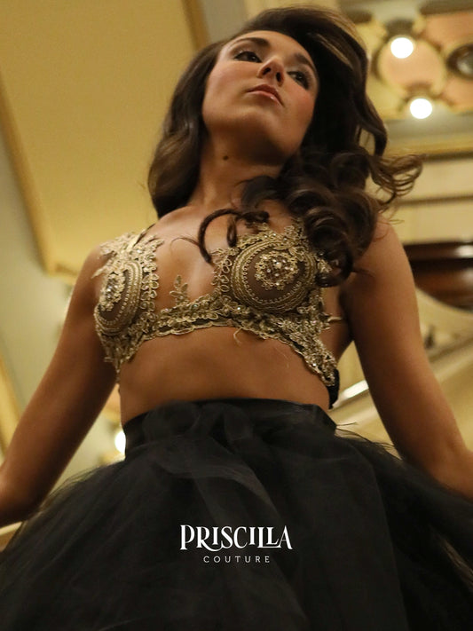 A woman wearing a black tulle ballgown with an ornate gold embellished bralette top, photographed in an indoor setting with warm lighting. The brand name 'PRISCILLA COUTURE' is printed on the black fabric at the bottom.