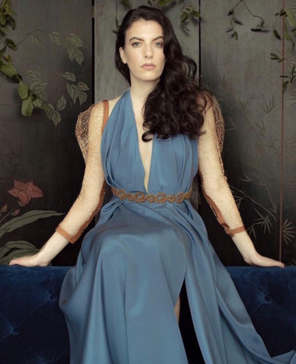 A woman sitting on a bench wearing a Priscilla Couture Romalter evening wear dress, blue, halter-neck, floor-length dress with a high-leg slit and gold detailing on the waist and sleeves.