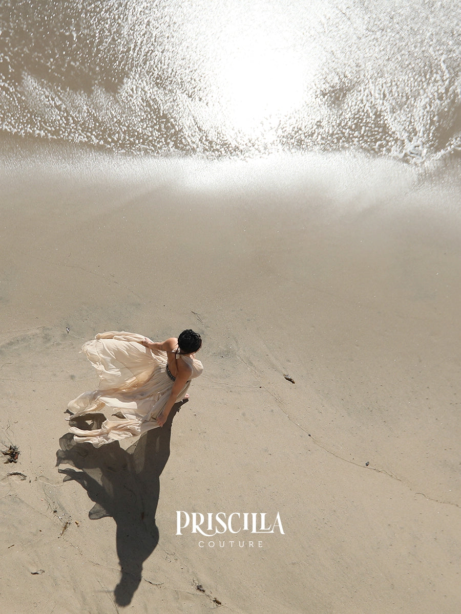 A woman in a flowing cream-colored dress stands on a sandy beach, viewed from above. The bright sun reflects off the wet sand and water near the top of the frame, and the brand name 'PRISCILLA COUTURE' is printed near the bottom.