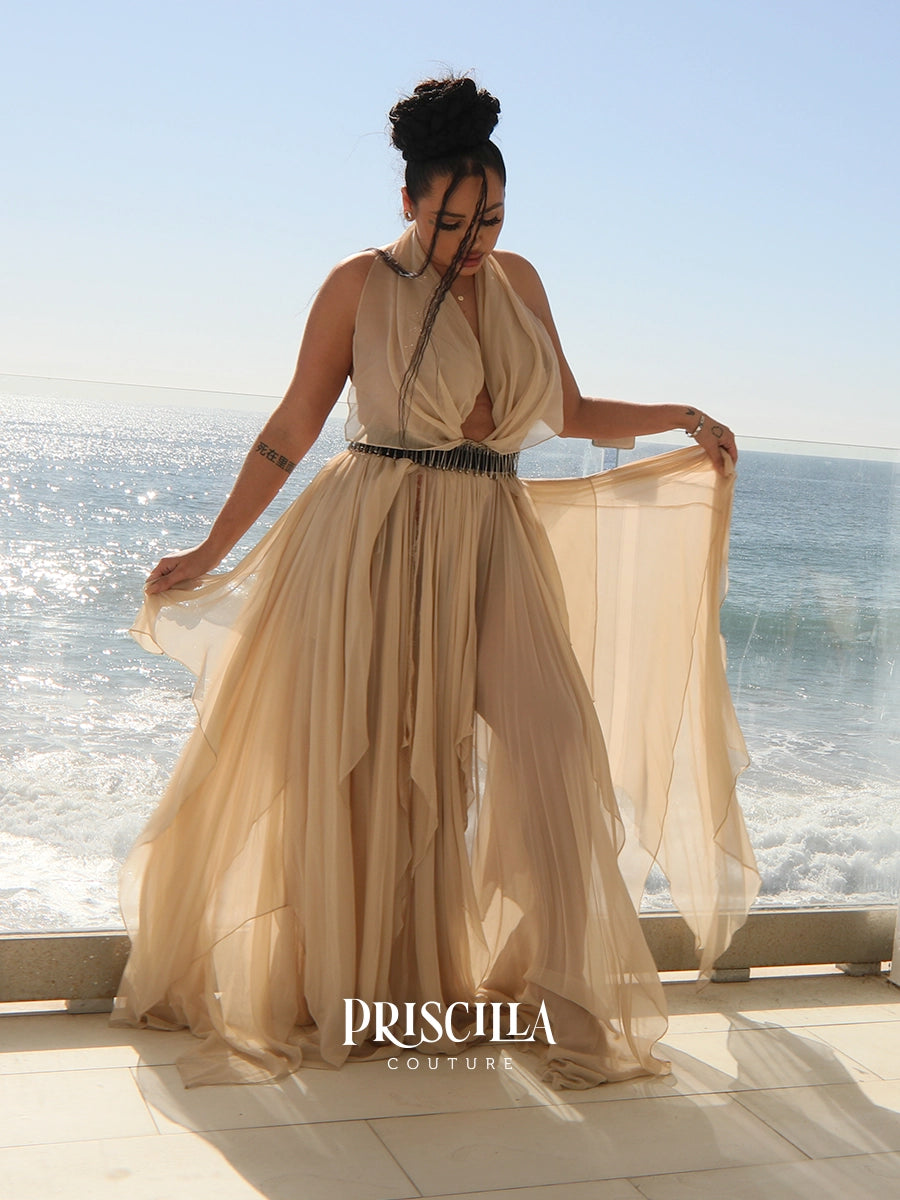 A woman wearing a flowing champagne beige halter maxi convertible skirt with a deep V-neckline and a thin black beaded waist belt, standing on a balcony with the ocean in the background. The brand name 'PRISCILLA COUTURE' is displayed at the bottom.