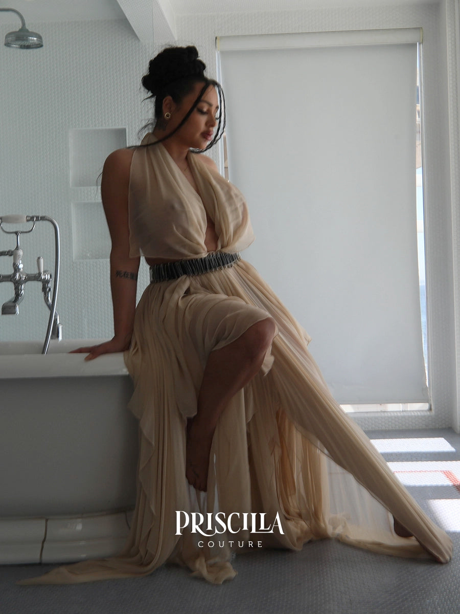 A woman models a flowing champagne beige convertible skirt with a deep V-neckline and a textured black beaded waistband, posing on the edge of a white bathtub in a modern bathroom. The brand name 'PRISCILLA COUTURE' is displayed at the bottom.