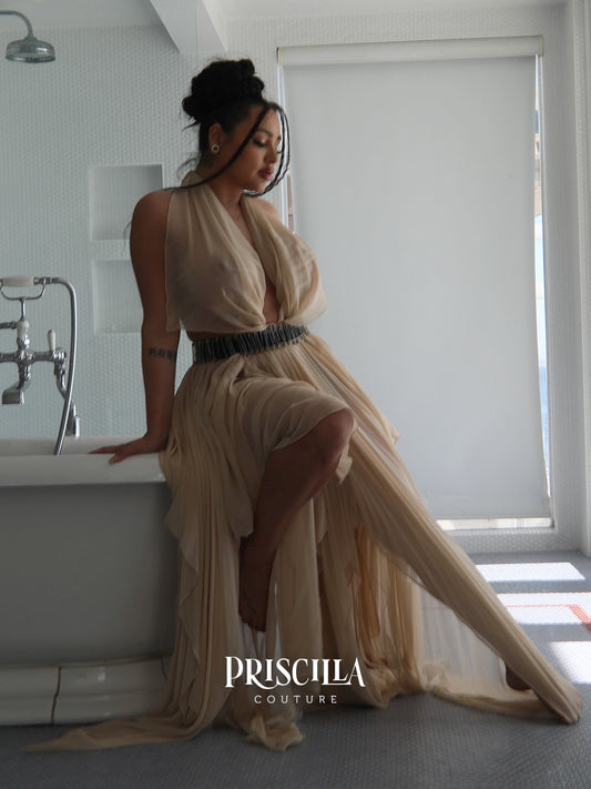 A woman models a flowing champagne beige convertible skirt with a deep V-neckline and a textured black beaded waistband, posing on the edge of a white bathtub in a modern bathroom. The brand name 'PRISCILLA COUTURE' is displayed at the bottom.
