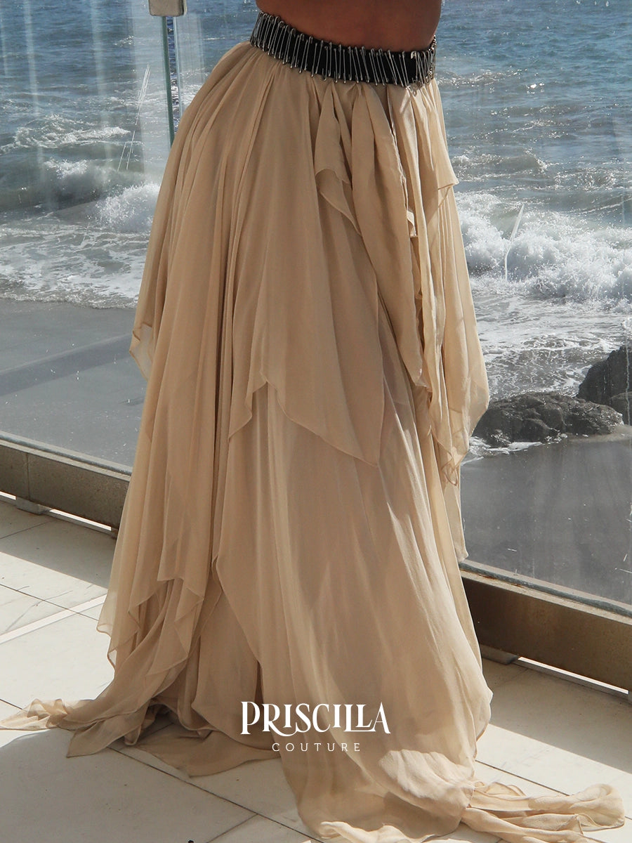 A beige flowy maxi skirt with a black beaded waistband, shown on a model standing on a balcony with the ocean in the background. The brand name 'PRISCILLA COUTURE' is visible at the bottom.