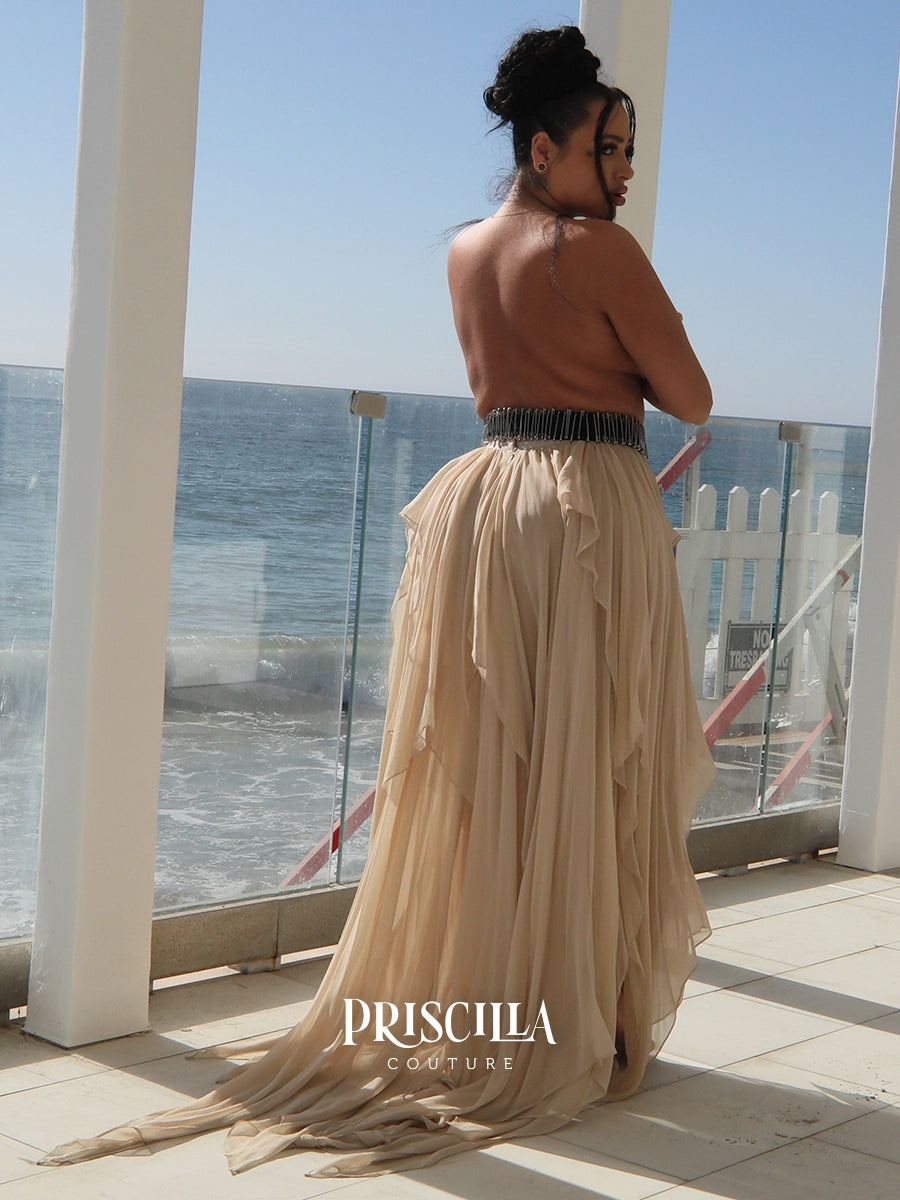 A woman stands on a sunlit balcony overlooking the ocean, wearing a strapless beige maxi convertible skirt with a silver safety pins waistband and a long flowing train. The brand name PRISCILLA COUTURE is printed on the dress.