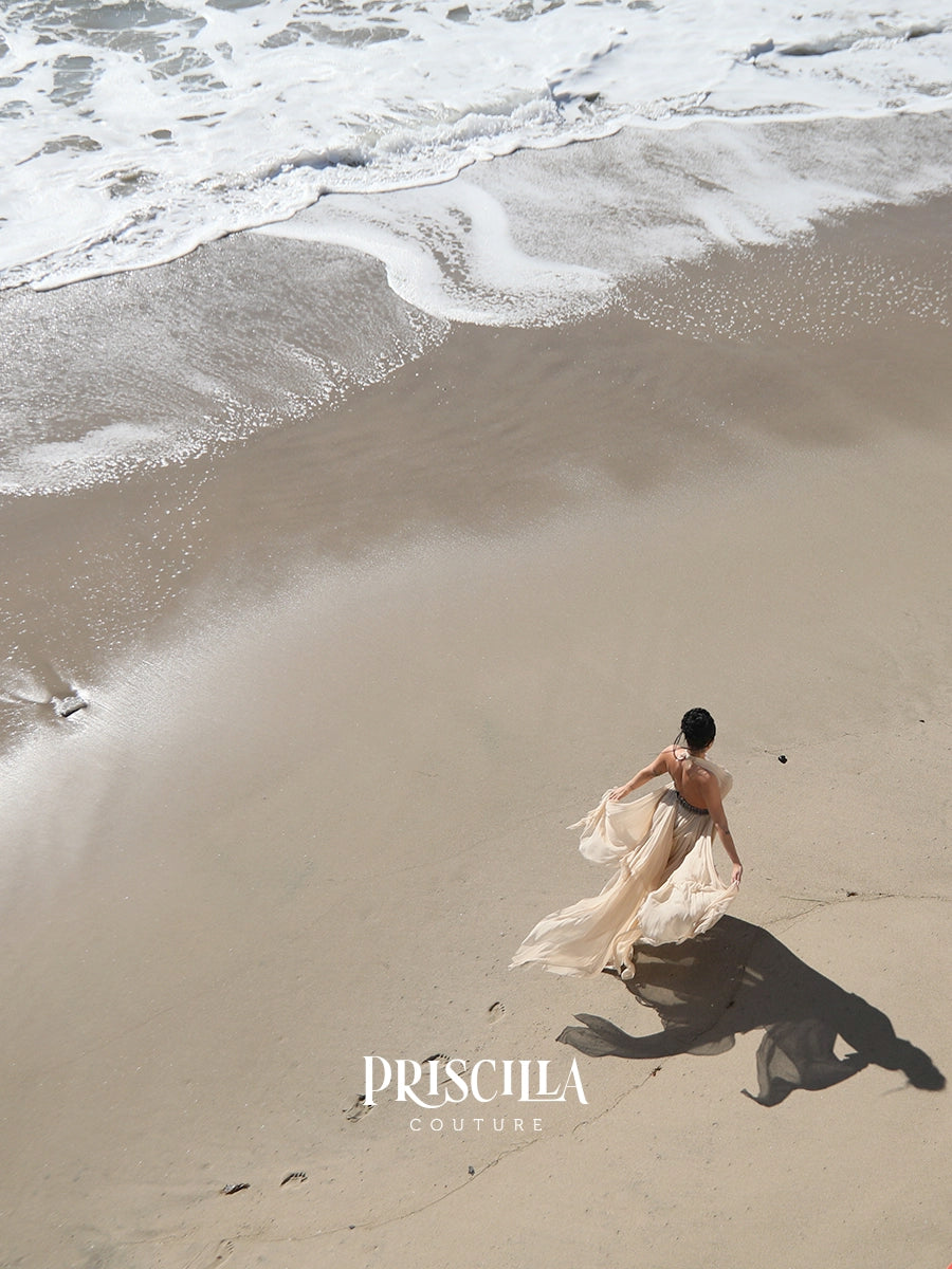 A woman in a flowing light beige convertible skirt walks on a sandy beach with ocean waves in the background. The brand name 'PRISCILLA COUTURE' is printed on the sand.
