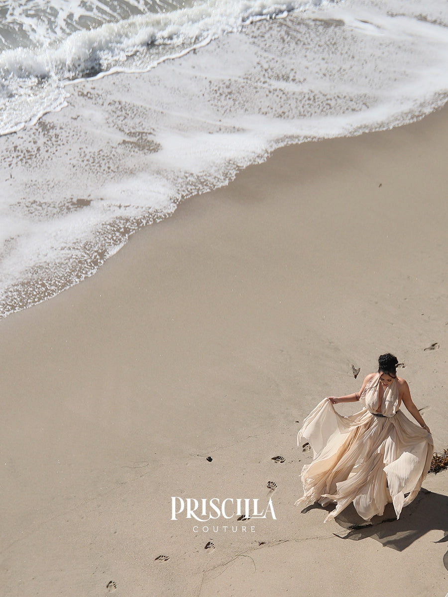 A woman in a flowing champagne-colored convertible wedding dress walks on a sandy beach with ocean waves in the background. The brand name 'PRISCILLA COUTURE' is displayed on the sand.