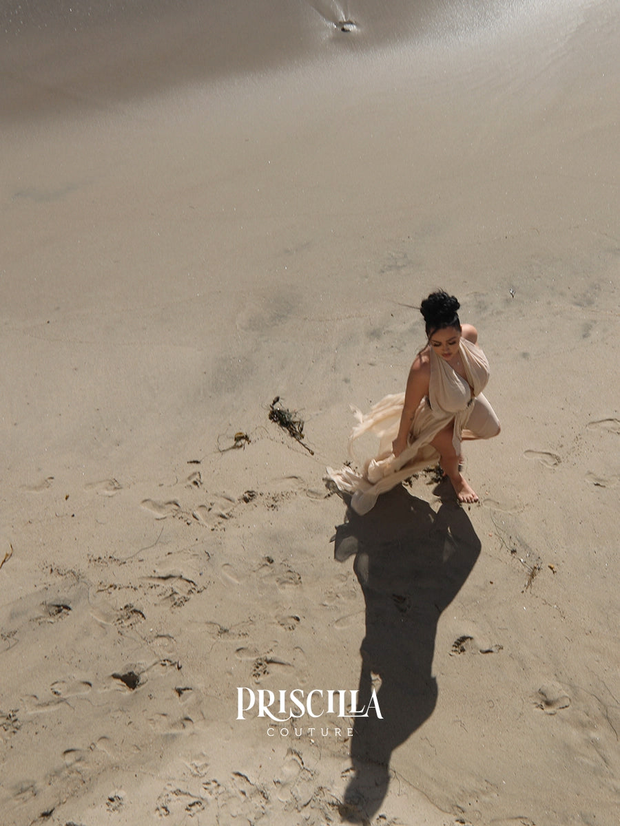 A woman in a flowing light beige convertible skirt crouches on a sandy beach, with her long shadow stretching across the sand and the brand name 'PRISCILLA COUTURE' printed near the bottom.