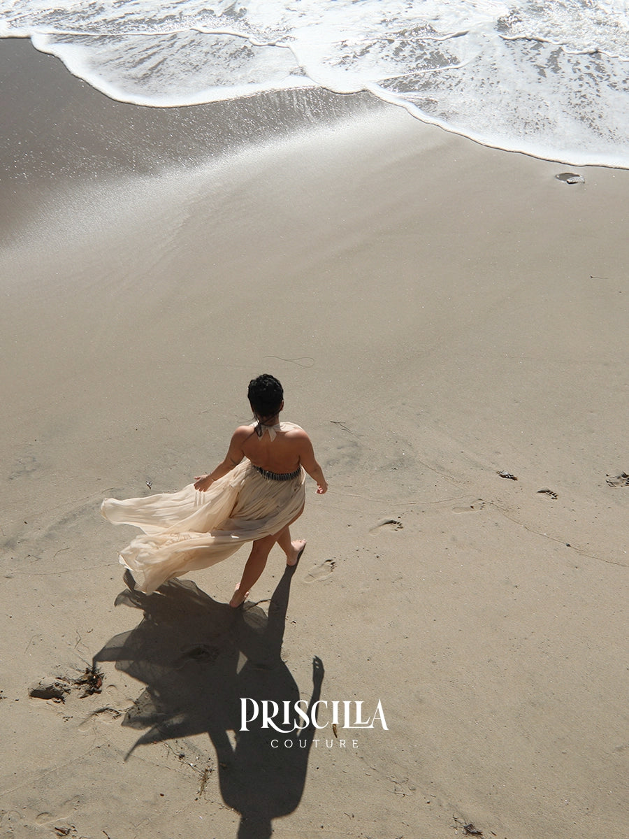 A woman stands on a sandy beach facing the ocean, wearing a flowing light beige Priscilla Couture convertible dress. The dress is caught in the wind and billows across the sand, casting a long shadow.