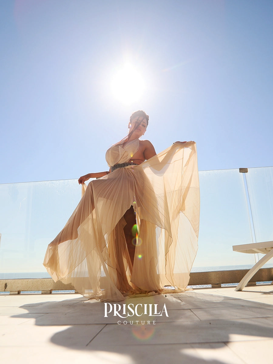 A woman wearing a flowing champagne beige halter-neck maxi convertible dress with a high leg slit, posing on a sunny outdoor terrace with the ocean and glass railing in the background. The brand name 'PRISCILLA COUTURE' is watermarked at the bottom.