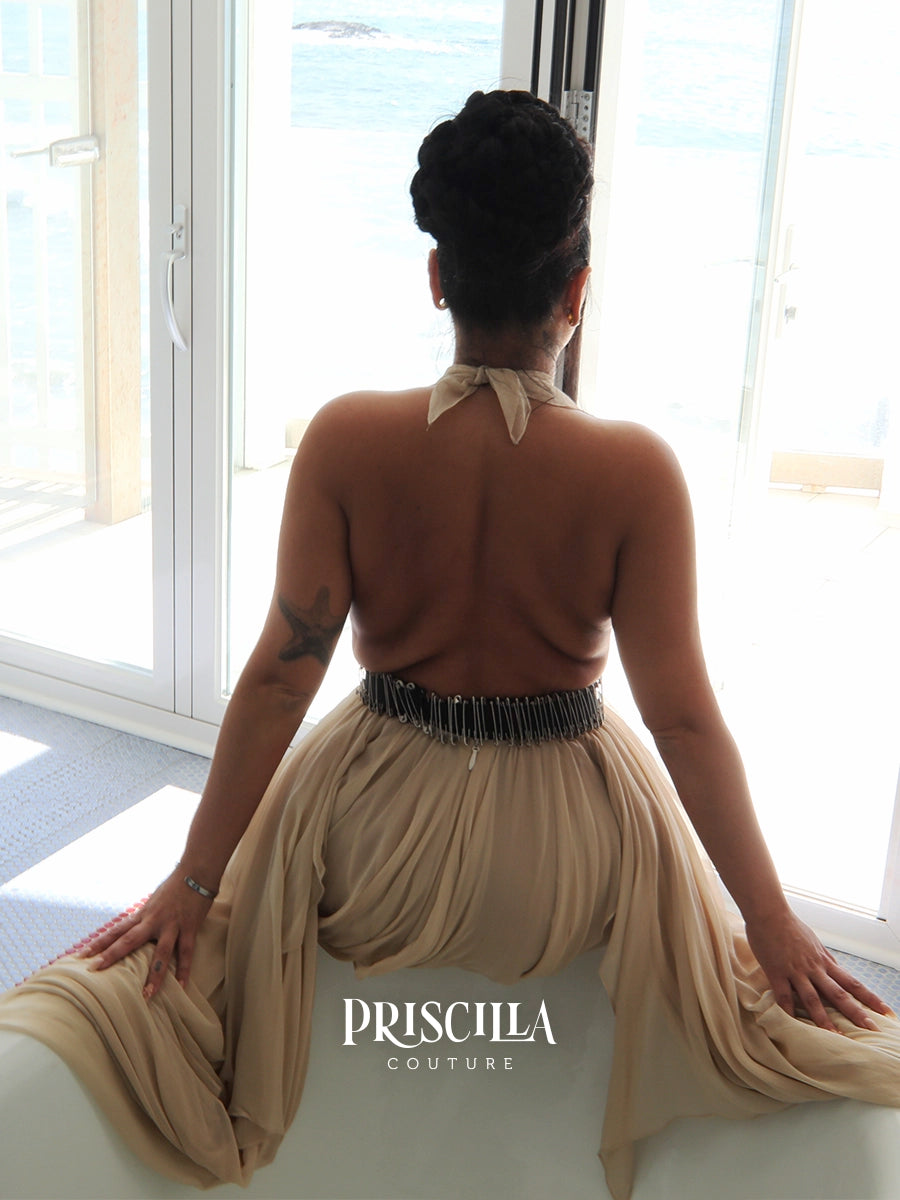 A woman is photographed from behind wearing a beige halter-neck draped convertible dress with a safety pins black waistband, sitting on a white bathtub in front of large windows overlooking water. The brand name 'PRISCILLA COUTURE' is printed on the image.