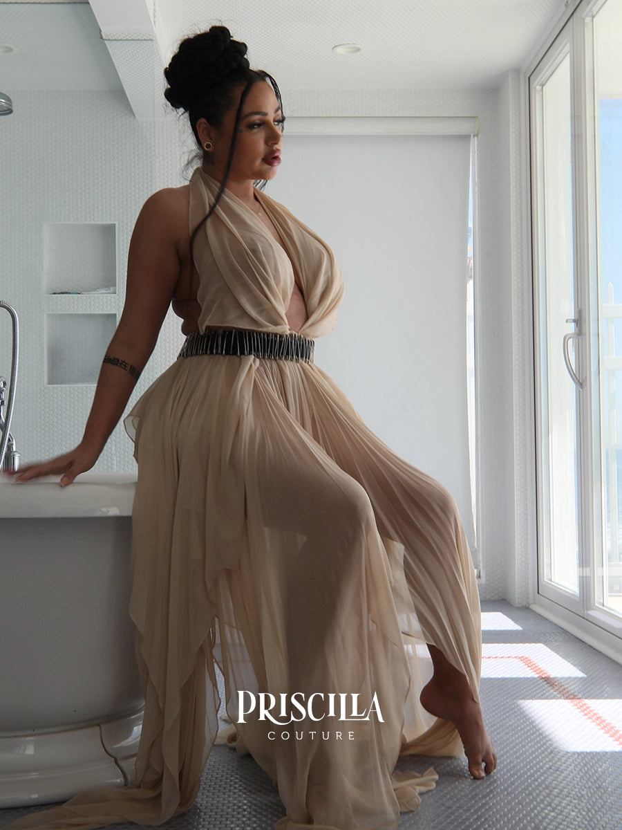 A woman models a beige halter-neck maxi convertible skirt with a deep V-neckline and a wide black embellished belt, posed in a bright, modern bathroom setting. The brand name 'PRISCILLA COUTURE' is watermarked at the bottom.