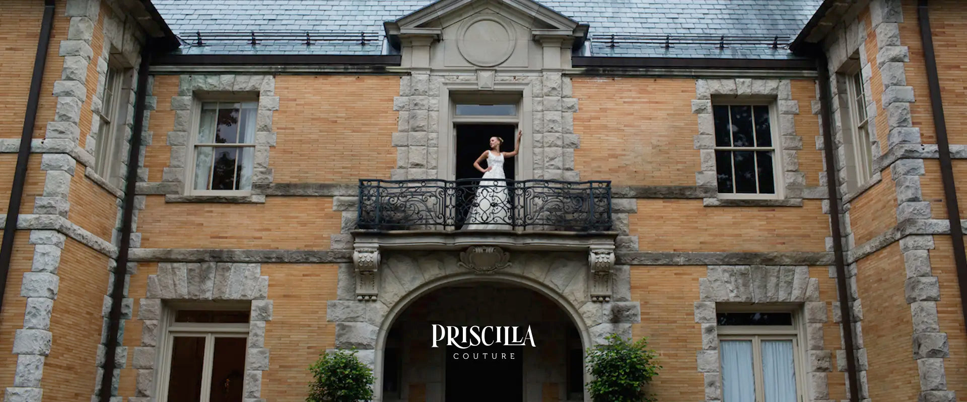 Bride in a white Priscilla Couture wedding dress standing on a balcony of a historic building with 'Priscilla Couture' branding.