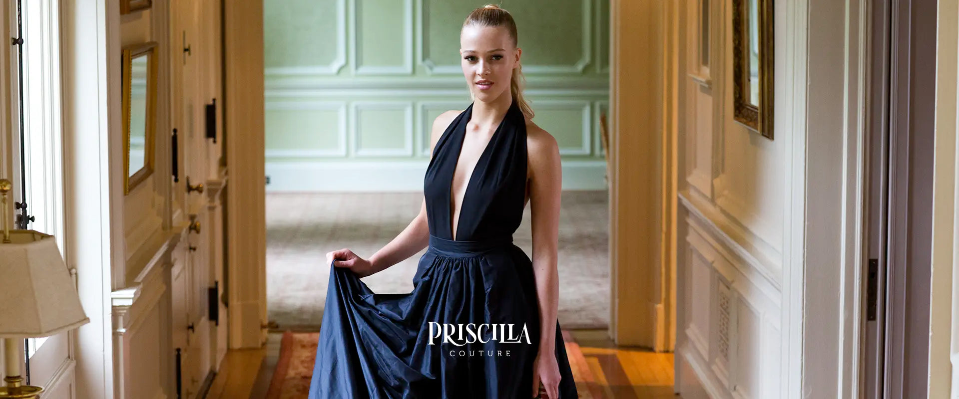 Woman in a Priscilla Couture's black top and black silk skirt standing in an elegant interior setting with 'Priscilla Couture' branding.