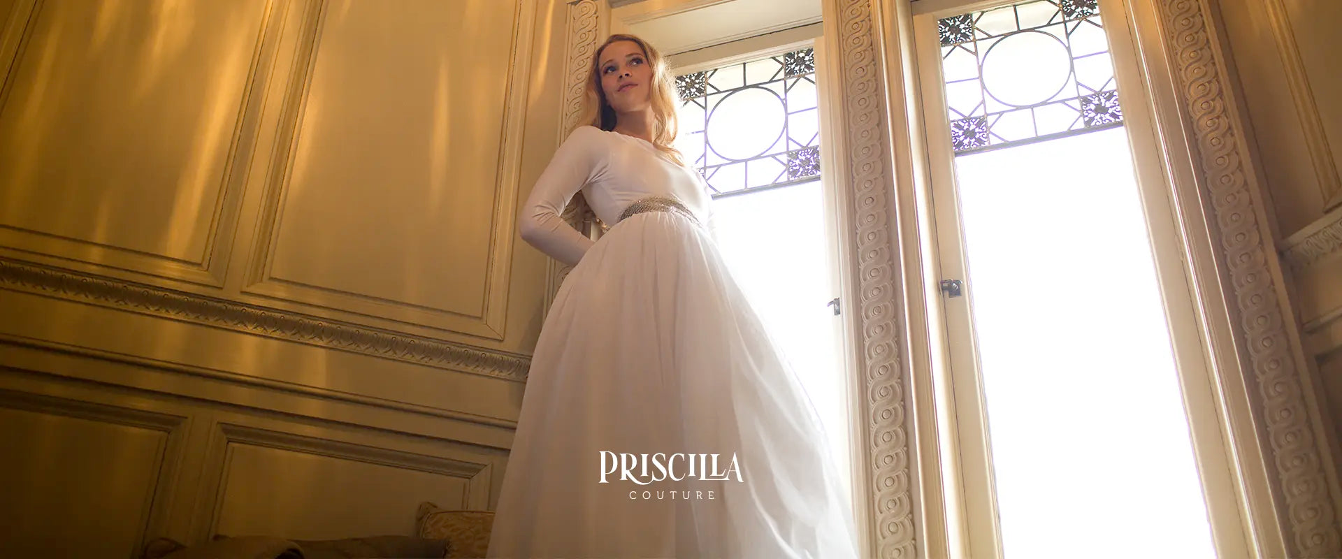 Bride in a  Priscilla Couture white wedding skirt standing in a room with decorative walls and stained glass windows, Priscilla Couture branding visible.