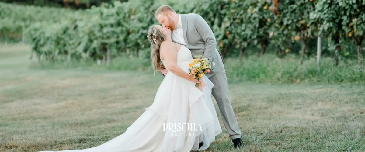 Bride in a Priscilla Couture custom wedding gown and her groom embracing in a vineyard with 'Priscilla Couture' branding. 