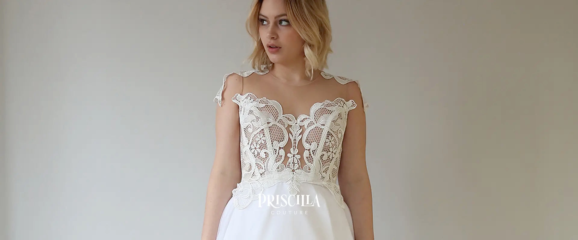 Bride wearing a white lace wedding dress with 'Priscilla Couture' branding against a plain background