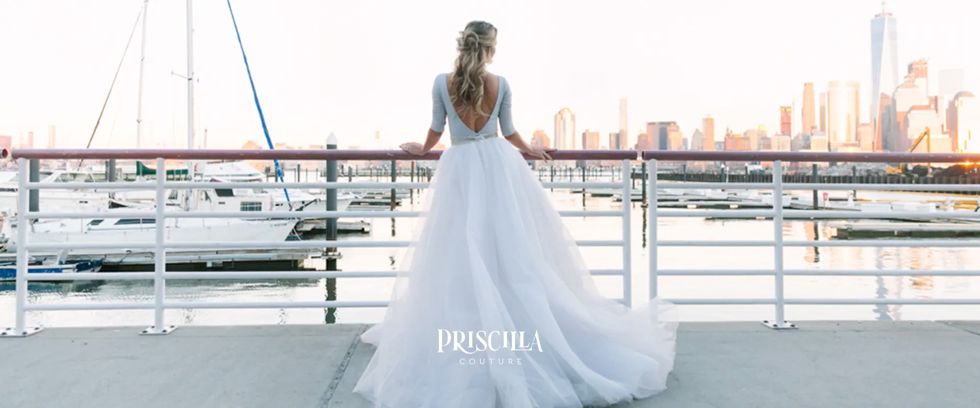 Bride in a Priscilla Couture white wedding tulle skirt standing on a dock with a city skyline in the background, Priscilla Couture brand visible.