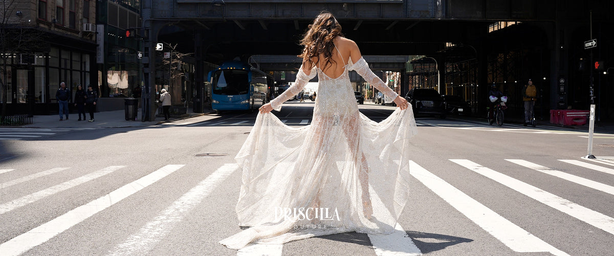 A fashion models stands on a NYC crosswalk wearing a Priscilla Couture crystal beaded sheer white off-the-shoulder wedding dress with long sleeves and a flowing train, holding the fabric out to the sides in an urban street setting.