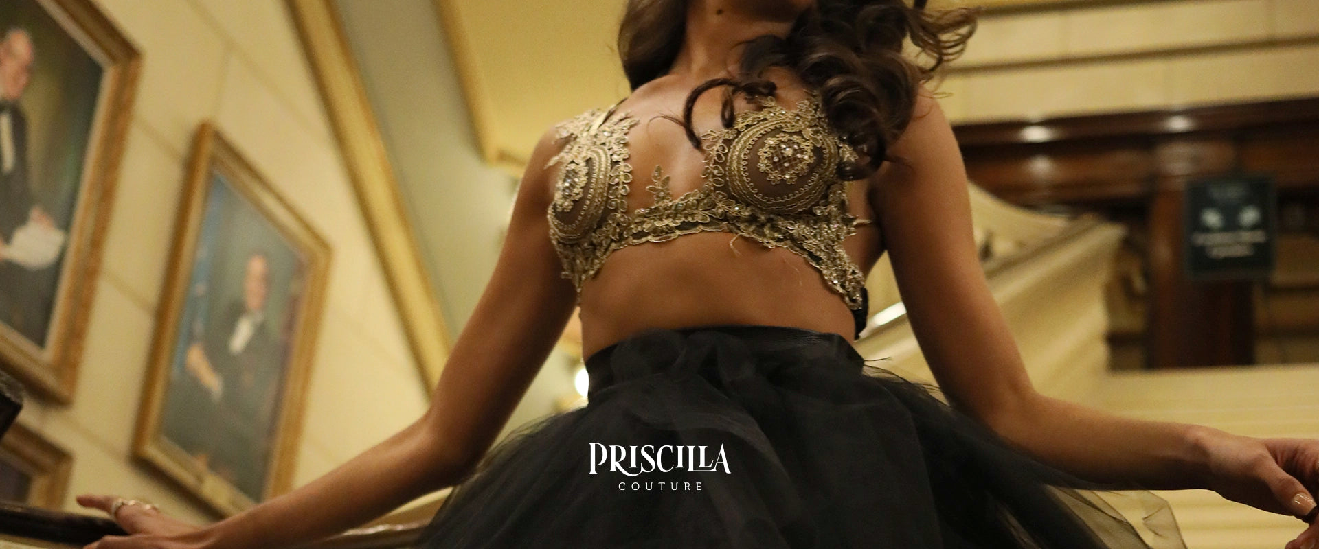 A model wearing a Priscilla Couture black and gold embellished bralette, photographed from a low angle in an indoor setting with warm lighting.